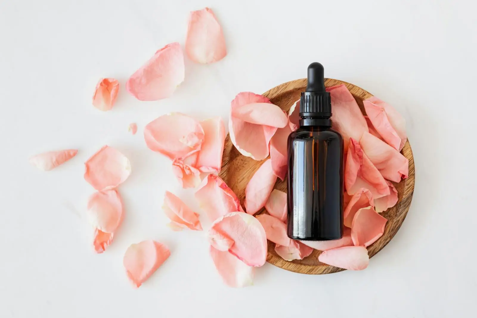 Top view of empty brown bottle for skin care product placed on wooden plate with fresh pink rose petals on white background isolated - natural facial moisturizers