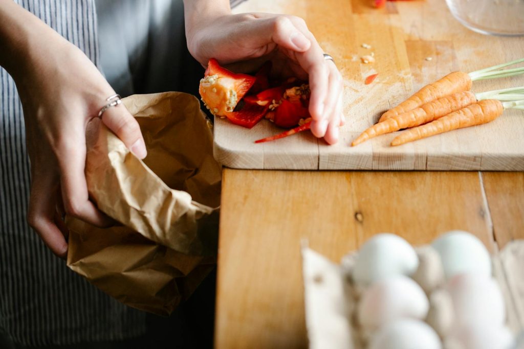 Unrecognizable female putting seeds of red bell pepper from cutting board into carton package while cooking at counter with carrots and eggs - Eco-Friendly Activities